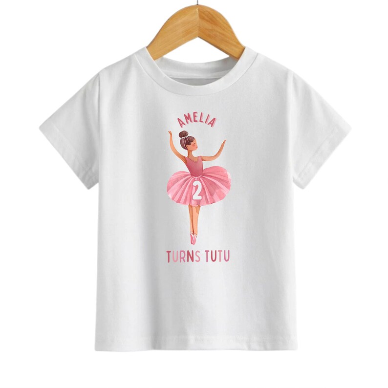 May include: White t-shirt with a pink ballerina graphic and the text "Amelia Turns Tutu" with the number 2 on the ballerina's skirt.