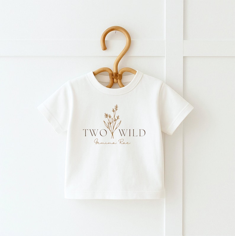 Puede incluir: Camiseta blanca con un dise&ntilde;o floral marr&oacute;n y el texto "TWO WILD" y "Gemima Rae" impreso en la parte delantera.