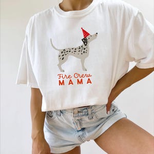 May include: White t-shirt with a Dalmatian dog illustration wearing a party hat. The dog has black spots and is standing. The text "Fire Crew MAMA" is printed in red. The shirt is a casual style, suitable for everyday wear.