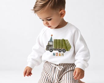 Puede incluir: Una sudadera blanca de manga larga con un camión de basura verde estilo acuarela, una pequeña estrella naranja en la parte superior y el nombre NOAH impreso en letras multicolores, junto a un pantalón corto de rayas verticales marrón y blanco.