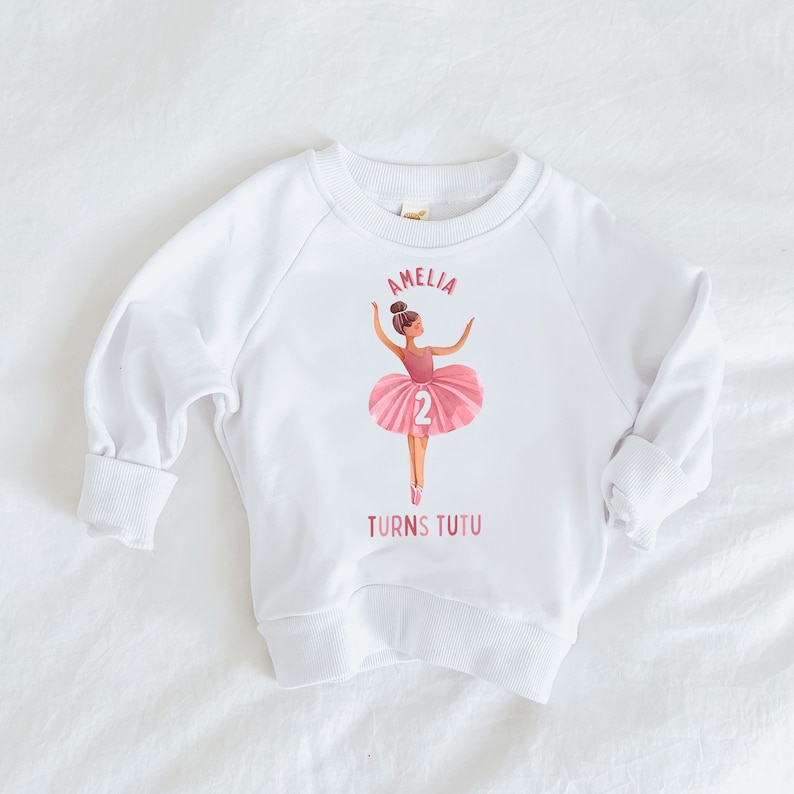 May include: White sweatshirt with a pink ballerina graphic and the text "Amelia turns tutu" and the number "2".