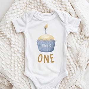 May include: White baby onesie with a blue and yellow cupcake graphic. The cupcake has a lit candle and the text "Finn's One" is printed below it.