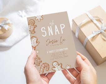 May include: A tan-colored invitation card with a gingerbread cookie theme. The text on the card reads "oh SNAP Josie Lu is turning two! A sweet celebration! Dec 12, 4 PM 1234 Cookie Way Christmas TX 123 456 7890".