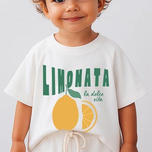 May include: White short-sleeved t-shirt featuring the word "LIMONATA" in green, a yellow lemon illustration, and the phrase "la dolce vita" in green. The shirt is worn by a child.