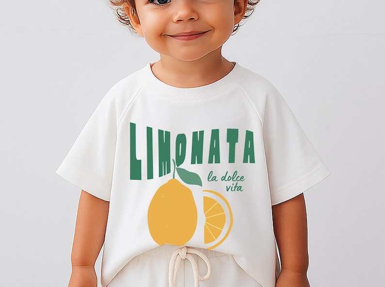 May include: White short-sleeved t-shirt featuring the word "LIMONATA" in green, a yellow lemon illustration, and the phrase "la dolce vita" in green. The shirt is worn by a child.