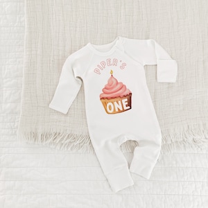 May include: White long-sleeve baby onesie with pink and yellow cupcake graphic and the text "Piper's One" in pink.