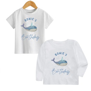 May include: Two white t-shirts featuring a blue whale wearing a party hat and the text "BOWIE'S Birthday". One t-shirt has short sleeves and the other has long sleeves.