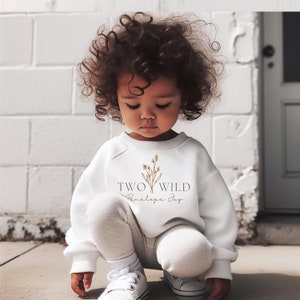 Puede incluir: Una sudadera blanca con un dise&ntilde;o floral marr&oacute;n y el texto "TWO WILD Penelope Joy" impreso en la parte delantera.
