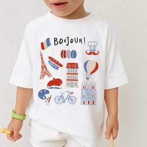 Op de afbeelding: Wit T-shirt met een kleurrijke Franse thema-afbeelding. Het ontwerp bevat de Eiffeltoren, een baguette, een baret, een luchtballon en de tekst "BONJOUR!" in blauw en rood. Het shirt is een model met korte mouwen.