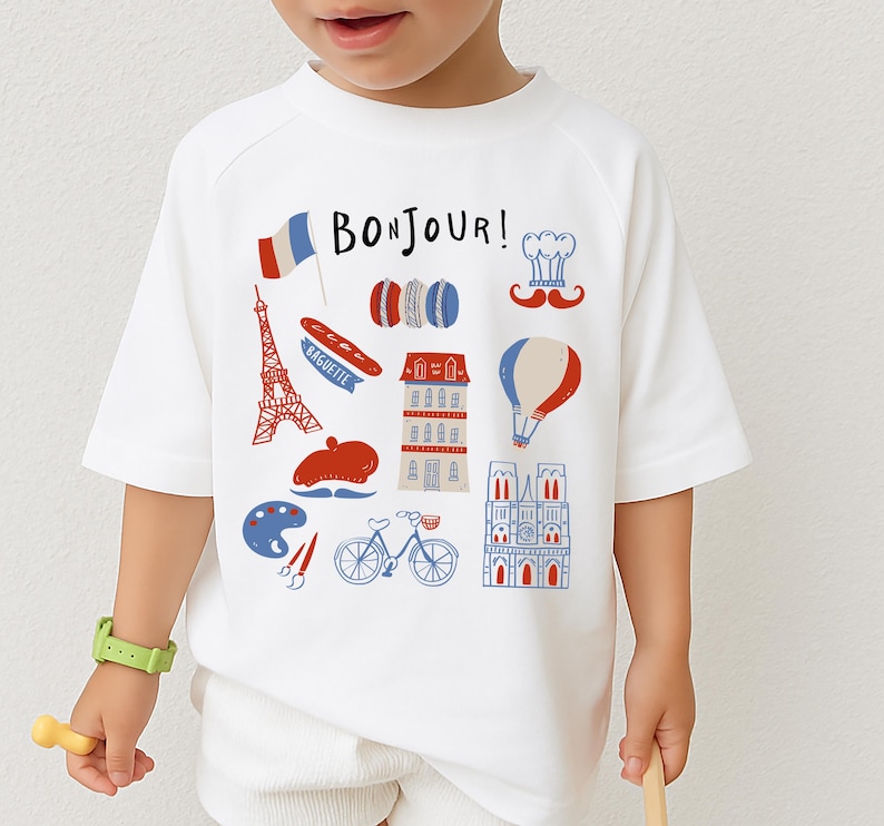 Op de afbeelding: Wit T-shirt met een kleurrijke Franse thema-afbeelding. Het ontwerp bevat de Eiffeltoren, een baguette, een baret, een luchtballon en de tekst "BONJOUR!" in blauw en rood. Het shirt is een model met korte mouwen.