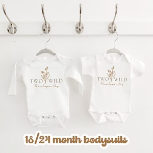 Puede incluir: Dos bodies de beb&eacute; blancos con mangas largas y cortas. Los bodies tienen un gr&aacute;fico marr&oacute;n de un dise&ntilde;o floral con el texto "TWO WILD Penelope Joy". Los bodies est&aacute;n colgados en perchas. El texto "18/24 month bodysuits" est&aacute; escrito en marr&oacute;n en la parte inferior de la imagen.