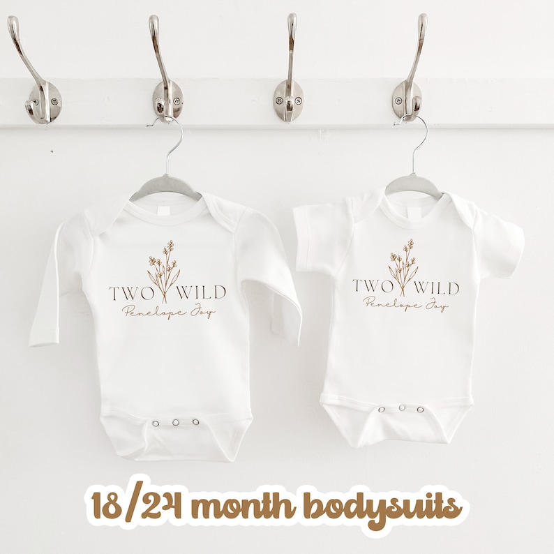 Puede incluir: Dos bodies de beb&eacute; blancos con mangas largas y cortas. Los bodies tienen un gr&aacute;fico marr&oacute;n de un dise&ntilde;o floral con el texto "TWO WILD Penelope Joy". Los bodies est&aacute;n colgados en perchas. El texto "18/24 month bodysuits" est&aacute; escrito en marr&oacute;n en la parte inferior de la imagen.