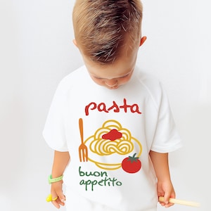 Op de afbeelding: Wit T-shirt met korte mouwen met een pasta-thema. Het ontwerp bevat het woord "pasta" in het rood, een bord spaghetti, een vork, een tomaat en de woorden "buon appetito" in het groen.