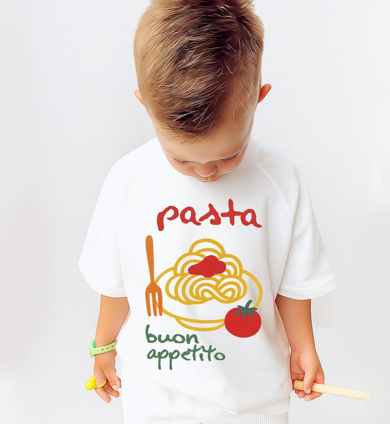 Op de afbeelding: Wit T-shirt met korte mouwen met een pasta-thema. Het ontwerp bevat het woord "pasta" in het rood, een bord spaghetti, een vork, een tomaat en de woorden "buon appetito" in het groen.