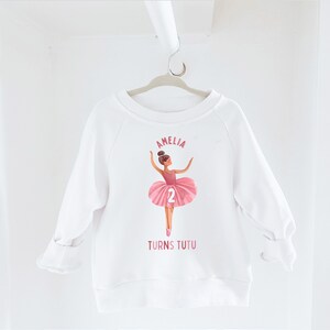 May include: White sweatshirt with a pink ballerina graphic and the text "Amelia Turns Tutu" and the number "2".