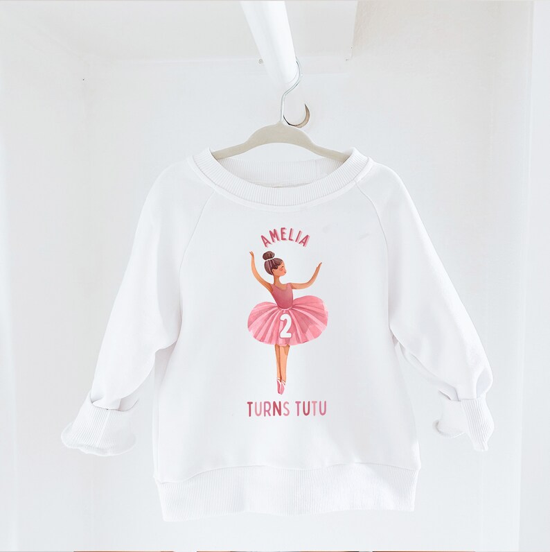 May include: White sweatshirt with a pink ballerina graphic and the text "Amelia Turns Tutu" and the number "2".
