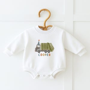 May include: White long-sleeved baby onesie with a green and brown garbage truck graphic and the name "COOPER" in colorful letters.