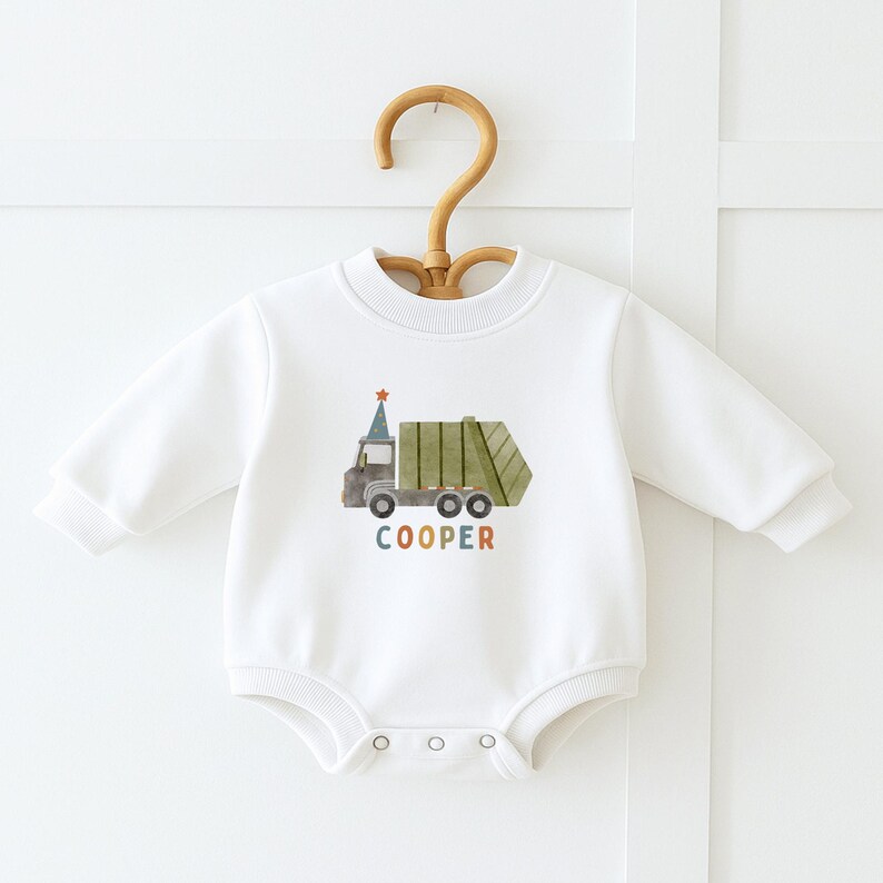 May include: White long-sleeved baby onesie with a green and brown garbage truck graphic and the name "COOPER" in colorful letters.