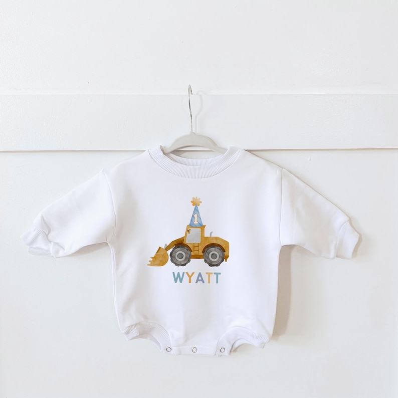 May include: White long-sleeved onesie with a yellow construction vehicle wearing a blue party hat with the number one on it. The text "WYATT" is printed below the vehicle.