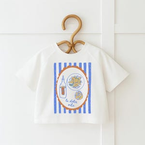 May include: White short-sleeved t-shirt with a graphic of Italian food. The design includes a bottle, pasta, and a lemon inside an oval frame with the text "la dolce vita". Blue vertical stripes are on the shirt.