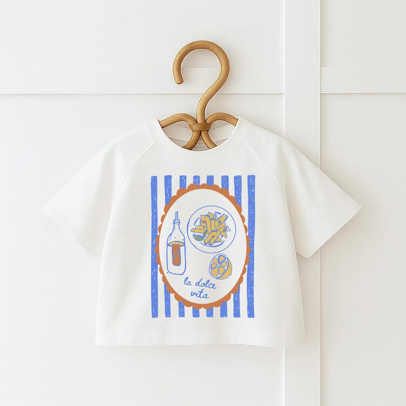 May include: White short-sleeved t-shirt with a graphic of Italian food. The design includes a bottle, pasta, and a lemon inside an oval frame with the text "la dolce vita". Blue vertical stripes are on the shirt.