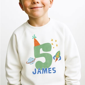 May include: A cream-colored long-sleeve sweatshirt featuring a large green number five wearing an orange party hat. The design includes a blue planet with rings, a colorful rocket ship, yellow stars, and the name JAMES printed in blue letters.