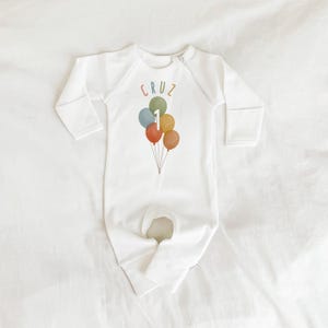 Może przedstawiać: Biały, długorękawny śpioszek dla niemowlęcia z zamkiem błyskawicznym. Śpioszek ma kolorową grafikę z balonami z numerem "1" i imieniem "CRUZ" nadrukowanym z przodu.