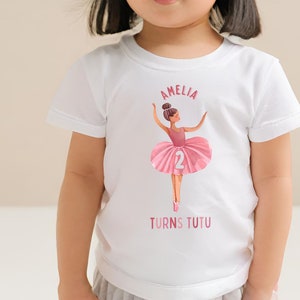 May include: White t-shirt with a pink ballerina illustration and the text "Amelia Turns Tutu" and the number "2".