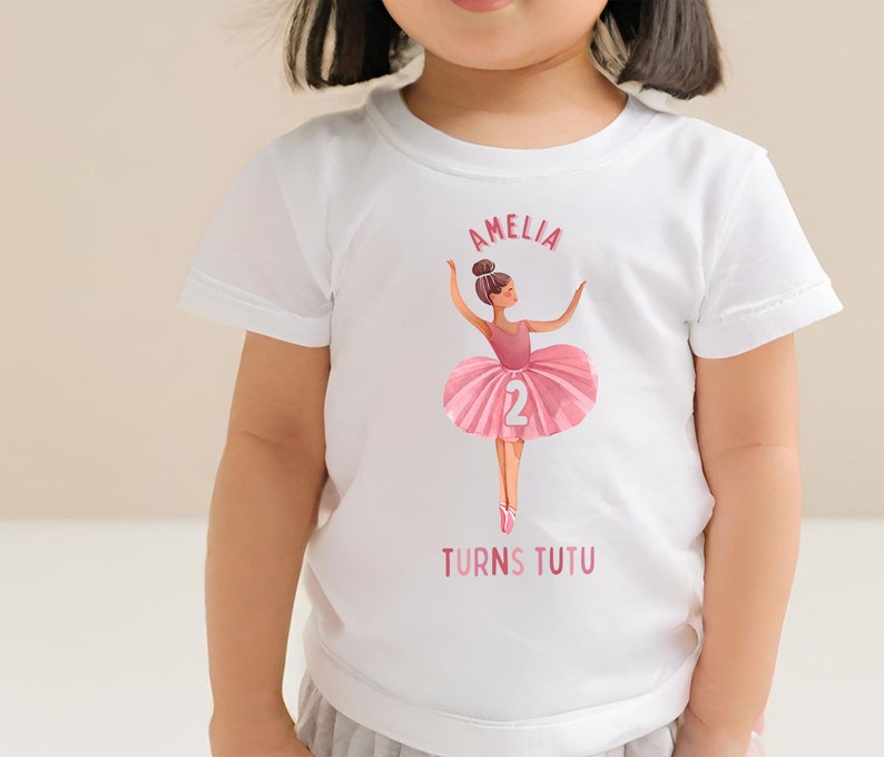 May include: White t-shirt with a pink ballerina illustration and the text "Amelia Turns Tutu" and the number "2".