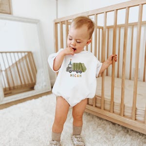 May include: A white baby onesie with a green garbage truck graphic and the name Micah in colorful letters. The baby is wearing the onesie and standing next to a wooden crib.