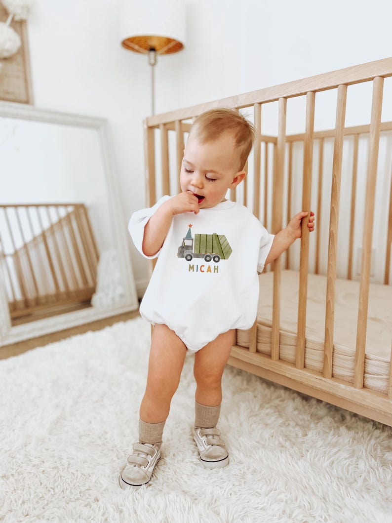 May include: A white baby onesie with a green garbage truck graphic and the name Micah in colorful letters. The baby is wearing the onesie and standing next to a wooden crib.