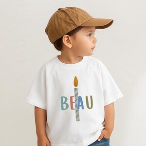 Pode incluir: Uma crian&ccedil;a usando um bon&eacute; de beisebol marrom e uma camiseta branca. A camiseta apresenta um gr&aacute;fico de vela colorida com o nome "BEAU" em letras azuis, rosa e verdes. A crian&ccedil;a est&aacute; usando jeans azuis.