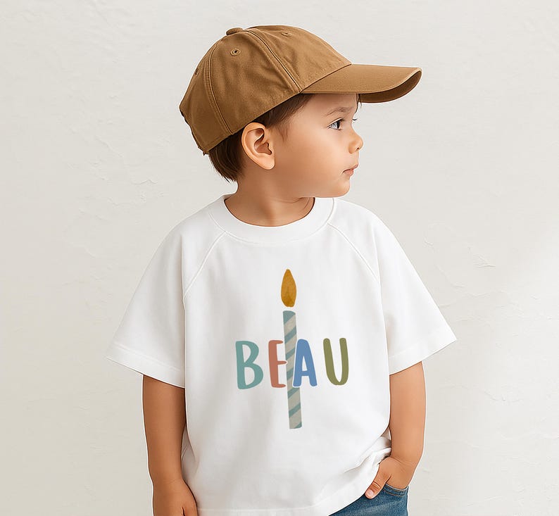 Pode incluir: Uma crian&ccedil;a usando um bon&eacute; de beisebol marrom e uma camiseta branca. A camiseta apresenta um gr&aacute;fico de vela colorida com o nome "BEAU" em letras azuis, rosa e verdes. A crian&ccedil;a est&aacute; usando jeans azuis.
