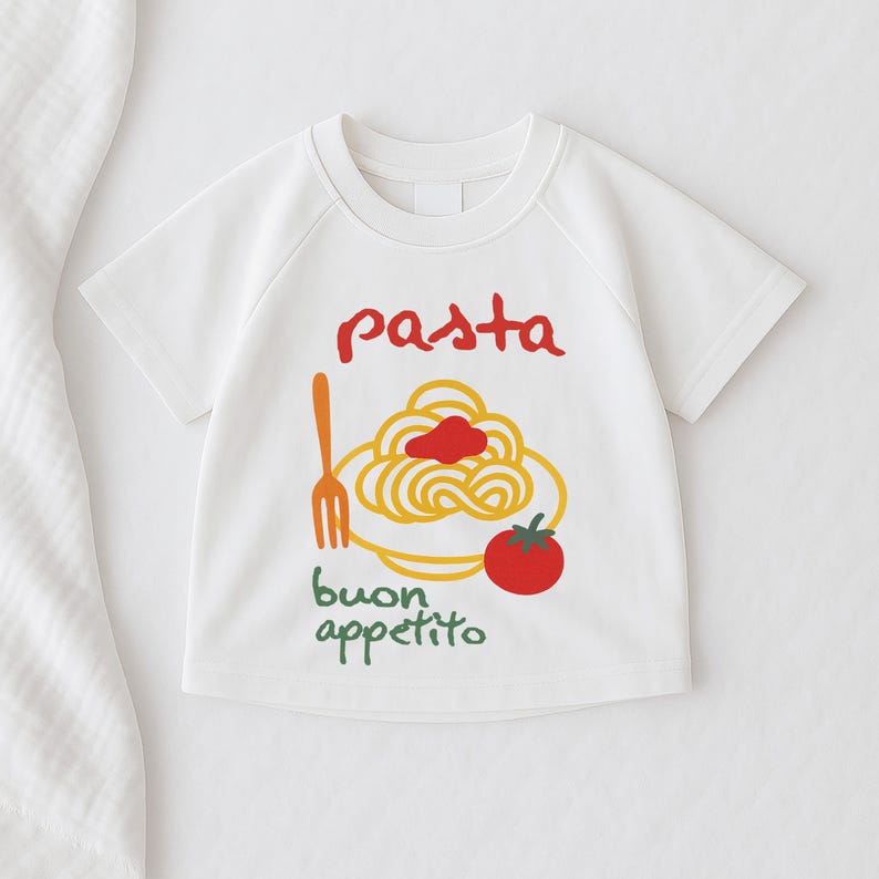 Op de afbeelding: Wit T-shirt met korte mouwen met een pasta-thema. Het ontwerp bevat het woord "pasta" in het rood, een bord spaghetti, een vork, een tomaat en de woorden "buon appetito" in het groen.
