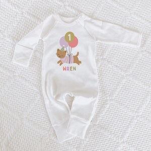 May include: White baby onesie with long sleeves. The onesie features a graphic of a brown cat wearing a party hat and holding balloons with the number 1. The name "WREN" is printed below the cat in colorful letters.