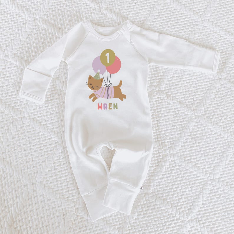 May include: White baby onesie with long sleeves. The onesie features a graphic of a brown cat wearing a party hat and holding balloons with the number 1. The name "WREN" is printed below the cat in colorful letters.