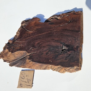 May include: A dark brown, irregularly shaped wood slab with a natural edge. The wood grain features swirling patterns and a knot. A small tag with the text "#160 1/2" is visible. The slab is on a white surface.