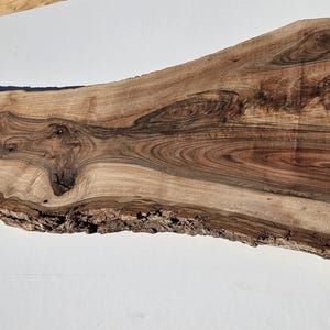 May include: A live edge wood slab with a natural, irregular shape. The slab features a mix of light and dark brown tones with intricate grain patterns. The bark edge is visible along the bottom. A small tag with text is in the lower left corner.