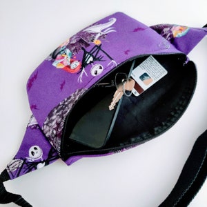 Może przedstawiać: Fioletowa nerka z czarnym paskiem i nadrukiem z filmu Koszmar przed świętami. Nerka jest otwarta i widać w niej czarny telefon i klucze.