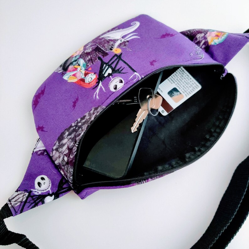 Może przedstawiać: Fioletowa nerka z czarnym paskiem i nadrukiem z filmu Koszmar przed świętami. Nerka jest otwarta i widać w niej czarny telefon i klucze.