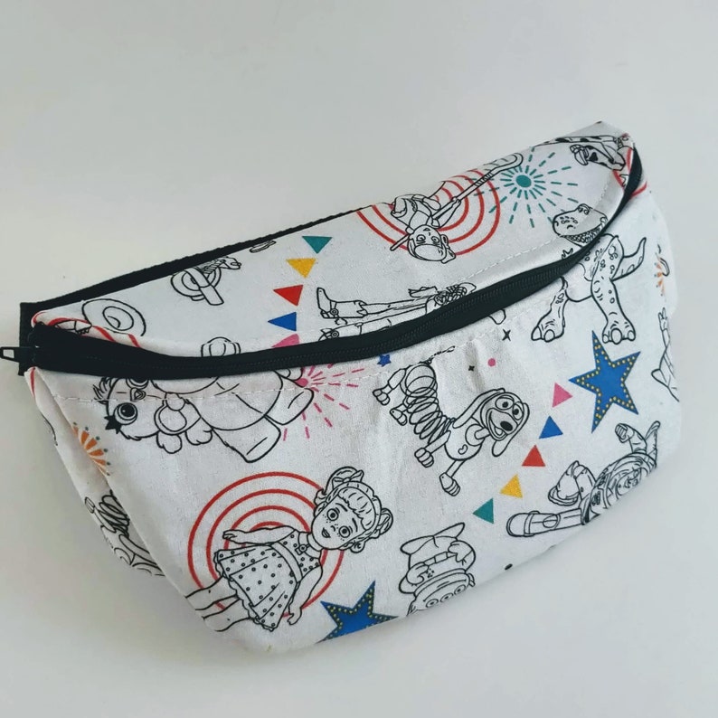 Puede incluir: Ri&ntilde;onera blanca con una cremallera negra y un estampado colorido de personajes de Toy Story. Los personajes est&aacute;n delineados en negro y rellenos de varios colores. El estampado incluye a Woody, Buzz Lightyear, Jessie, Slinky Dog, Rex y Hamm.