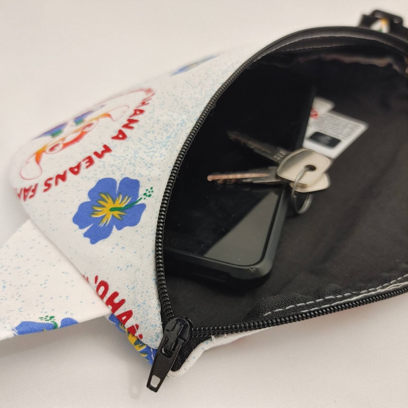 Puede incluir: Una ri&ntilde;onera blanca con cierre de cremallera. La ri&ntilde;onera tiene un estampado floral con flores azules y el texto "Aloha Means Family" en rojo y blanco.