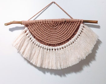 May include: A macrame wall hanging with a brown and white color scheme. The hanging is in the shape of a half circle with a fringe border. It is made with natural fibers and has a rustic, bohemian style.