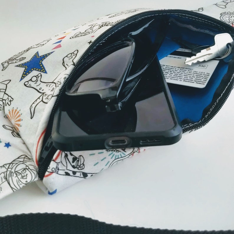 Puede incluir: Una ri&ntilde;onera de tela blanca con una cremallera negra y un estampado de dibujos animados de colores. La ri&ntilde;onera est&aacute; abierta y contiene un tel&eacute;fono m&oacute;vil negro, gafas de sol y una llave con una tarjeta azul que dice "Tarjeta no v&aacute;lida a menos que se active en EE. UU."