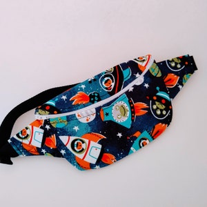 May include: A navy blue fanny pack with a white zipper and black strap. The pack is patterned with rockets, planets, stars, and aliens in orange, teal, and white. The bag is designed to be worn around the waist.