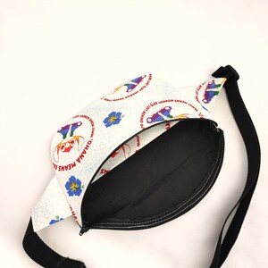 Puede incluir: Ri&ntilde;onera blanca con una correa negra. La parte delantera de la bolsa tiene un fondo blanco con un patr&oacute;n repetido de personajes de dibujos animados coloridos y el texto "'Ohana means family. Family means nobody gets left behind."