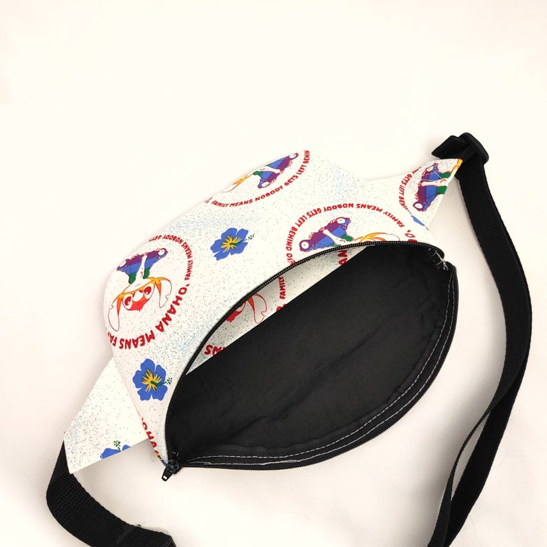 Puede incluir: Ri&ntilde;onera blanca con una correa negra. La parte delantera de la bolsa tiene un fondo blanco con un patr&oacute;n repetido de personajes de dibujos animados coloridos y el texto "'Ohana means family. Family means nobody gets left behind."