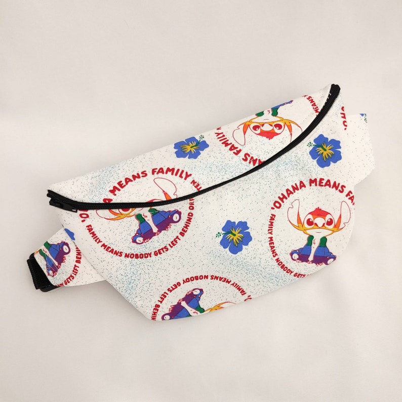 Puede incluir: Ri&ntilde;onera blanca con cremallera negra y un patr&oacute;n repetido de flores azules, personajes de dibujos animados rojos y amarillos, y el texto "Ohana Means Family" en rojo y negro.