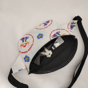 Puede incluir: Ri&ntilde;onera blanca con una correa negra y un interior blanco y negro. La parte delantera de la ri&ntilde;onera tiene un estampado colorido con el texto "Ohana Means Family" y "Family Means Nobody Gets Left Behind".