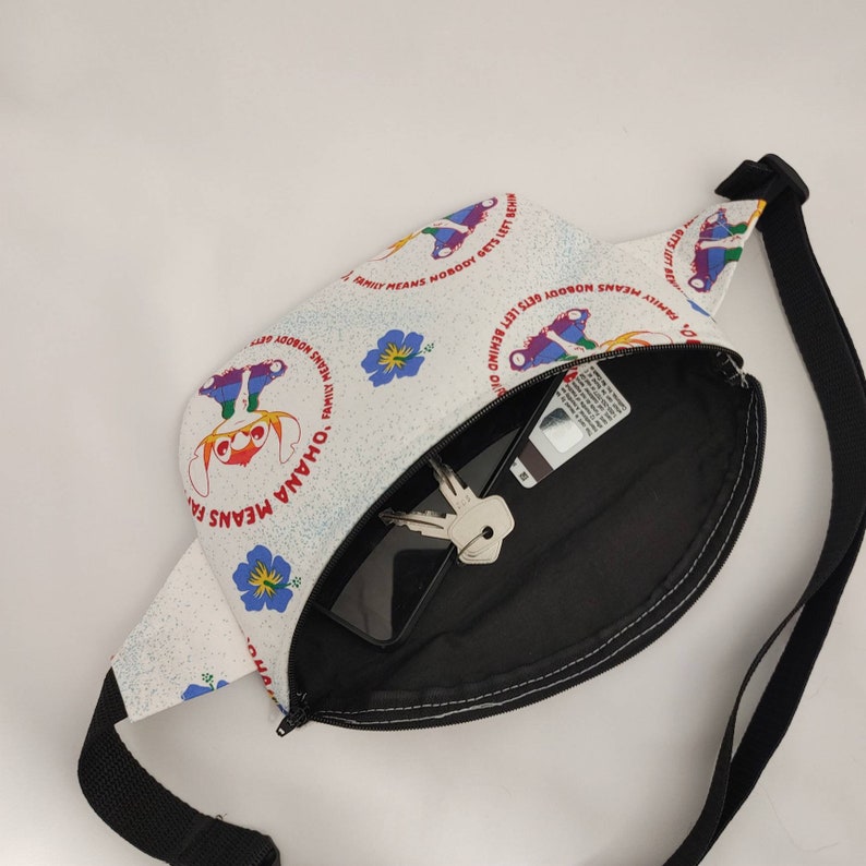 Puede incluir: Ri&ntilde;onera blanca con una correa negra y un interior blanco y negro. La parte delantera de la ri&ntilde;onera tiene un estampado colorido con el texto "Ohana Means Family" y "Family Means Nobody Gets Left Behind".